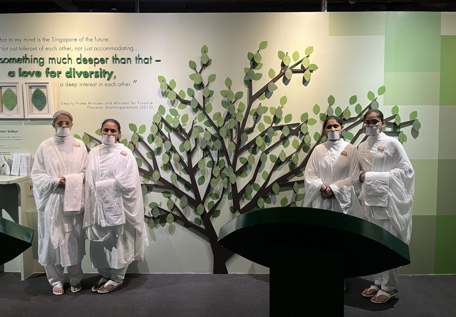 Four people in white robes with mouth covers stand before a wall mural of a tree with a diversity quote.